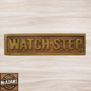 May include: A wooden sign with a brass frame and the words "WATCH STEP" in raised gold letters.