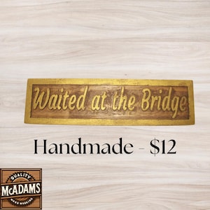 May include: A rectangular wooden sign with the words "Waited at the Bridge" in gold lettering. The sign has a gold border and is set against a light wood grain background. The word "Handmade" is below the sign.