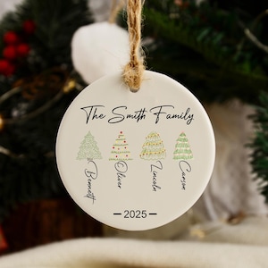 Può includere: Un ornamento natalizio rotondo in ceramica bianca con un cordino di iuta per appenderlo. L'ornamento presenta la scritta "The Smith Family" e quattro alberi di Natale illustrati, con nomi e l'anno 2025.