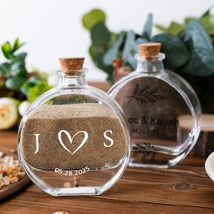 Pode incluir: Duas garrafas de vidro transparente para cerimônia de areia com rolhas de cortiça. Uma garrafa é preenchida com areia e apresenta as iniciais "J S" com um coração e a data "05.28.2025". A outra garrafa está vazia e tem texto e um desenho de folha.