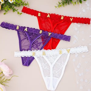 May include: Three thong underwear in red, purple, and white. Each thong has the word "BABY" spelled out in gold letters across the waistband. The red and purple thongs have a lace trim, while the white thong has a scalloped trim.