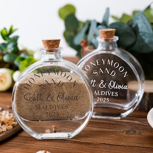 Pode incluir: Duas garrafas de vidro transparente com rolhas, cheias de areia. Uma garrafa é preenchida com areia e tem as palavras "Honeymoon Sand" e "Scott & Olivia Maldives 2025" impressas. A outra garrafa está vazia e tem o mesmo texto.