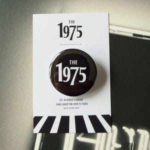 May include: A circular black pinback button featuring the white text THE 1975, mounted on a white display card. The card displays the text THE 1975 at the top and EST. IN DENISE'S GARAGE SAME LINEUP FOR OVER 23 YEARS at the bottom with black stripes.
