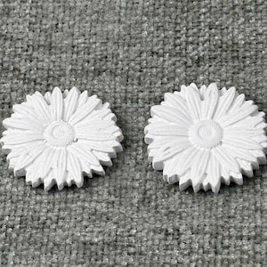 May include: Two white, daisy-shaped decorative objects. Each has a detailed center and numerous petals. The objects are made of a solid material and are placed on a textured, gray surface. The daisies are similar in size and shape.