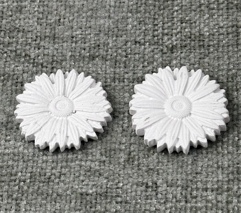 May include: Two white, daisy-shaped decorative objects. Each has a detailed center and numerous petals. The objects are made of a solid material and are placed on a textured, gray surface. The daisies are similar in size and shape.