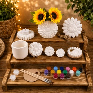 May include: A wooden tiered display with craft supplies. It features white flower-shaped objects, a vase with sunflowers, paint pots, paintbrushes, and a palette. The display is on a wooden surface.