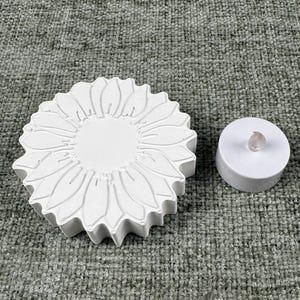 May include: A white, sunflower-shaped candle holder with detailed petal carvings. Beside it is a small, white, round LED candle with a clear, plastic flame. The items are on a textured, gray surface.