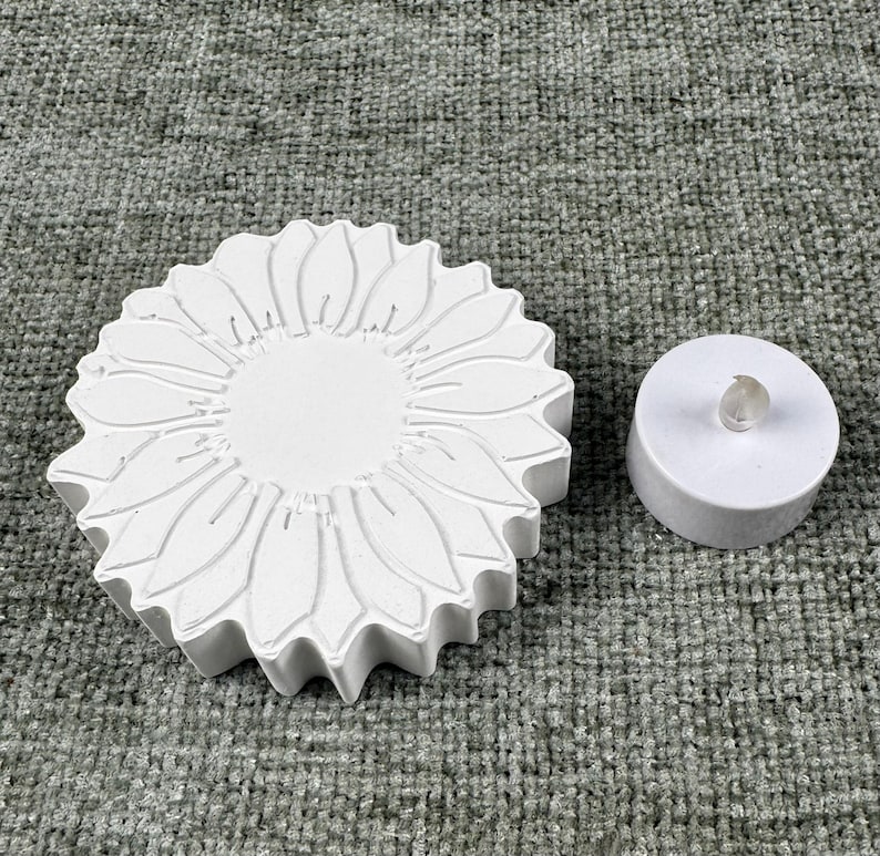 May include: A white, sunflower-shaped candle holder with detailed petal carvings. Beside it is a small, white, round LED candle with a clear, plastic flame. The items are on a textured, gray surface.