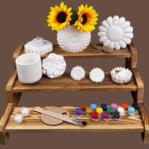 May include: A three-tiered wooden display shelf holds various craft items. It features a white vase with sunflowers, a candle holder, a small cup, and several sunflower-shaped decorations. The bottom shelf displays paint pots, a palette, and paintbrushes.