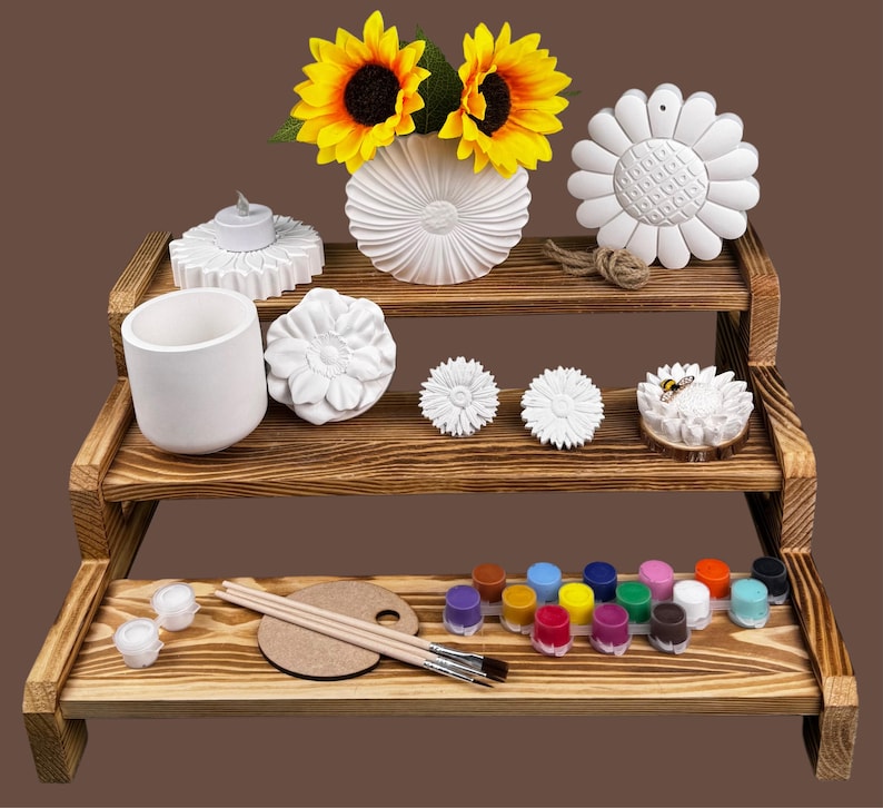 May include: A three-tiered wooden display shelf holds various craft items. It features a white vase with sunflowers, a candle holder, a small cup, and several sunflower-shaped decorations. The bottom shelf displays paint pots, a palette, and paintbrushes.
