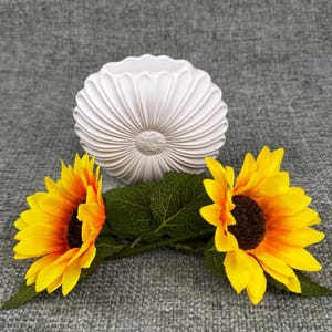 May include: A white, fluted ceramic vase with a sunflower design, accompanied by two sunflowers with yellow petals and dark brown centers. The sunflowers have green leaves and stems. The background is a gray fabric.