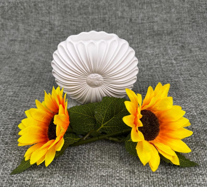 May include: A white, fluted ceramic vase with a sunflower design, accompanied by two sunflowers with yellow petals and dark brown centers. The sunflowers have green leaves and stems. The background is a gray fabric.