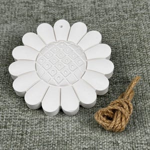 May include: A white, daisy-shaped ornament with a textured center and petal design. A small bundle of brown twine is next to the ornament. The ornament has a hole at the top for hanging.
