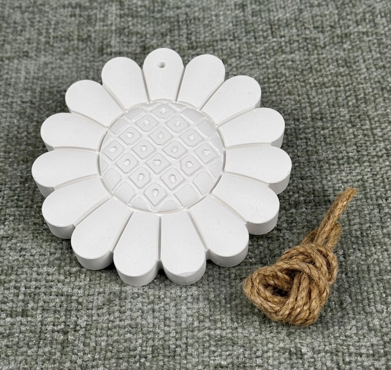 May include: A white, daisy-shaped ornament with a textured center and petal design. A small bundle of brown twine is next to the ornament. The ornament has a hole at the top for hanging.