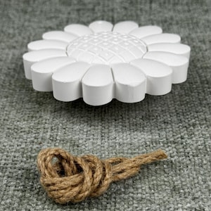 May include: A white, daisy-shaped decorative object with a textured center and rounded petals. Below it, a brown rope is tied in a decorative knot. The objects are set against a gray, textured background.