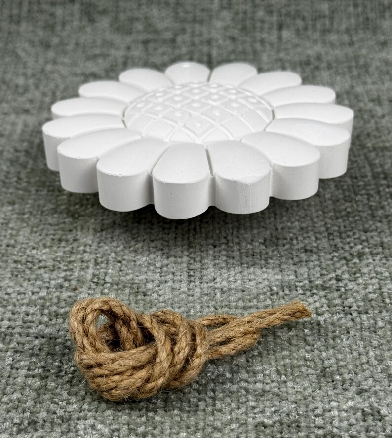 May include: A white, daisy-shaped decorative object with a textured center and rounded petals. Below it, a brown rope is tied in a decorative knot. The objects are set against a gray, textured background.