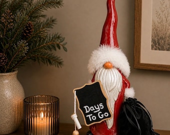 May include: A red and white gnome figurine with a chalkboard sign that reads "Days To Go." The gnome has a long red hat, a white beard, and a fluffy white collar. A black velvet bag sits next to the gnome. A lit candle is in a glass holder.