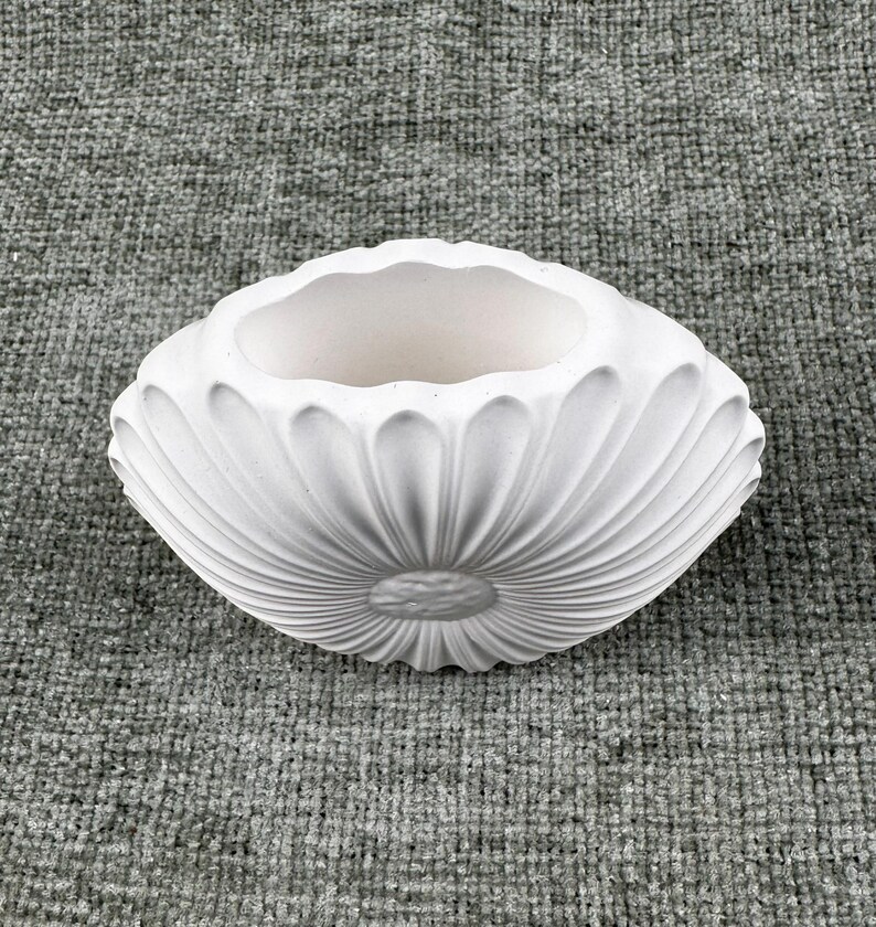 May include: A white, oval-shaped ceramic planter with a fluted, petal-like design. The interior is smooth and unadorned, contrasting with the textured exterior. The planter is suitable for small plants or decorative use.