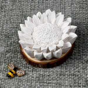 May include: A white sunflower-shaped decorative item on a wooden base, with a detailed textured center and petals. A small, gold-colored bee-shaped brooch with black and yellow stripes and rhinestone accents is next to the sunflower.