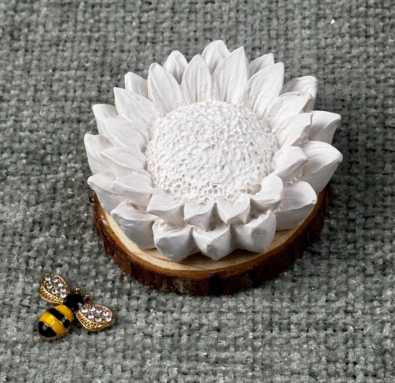 May include: A white sunflower-shaped decorative item on a wooden base, with a detailed textured center and petals. A small, gold-colored bee-shaped brooch with black and yellow stripes and rhinestone accents is next to the sunflower.