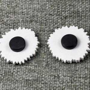 May include: Two white, gear-shaped magnets with a black, circular magnet in the center. The magnets are on a gray, textured surface. The gear-shaped design adds a unique touch to these functional magnets.