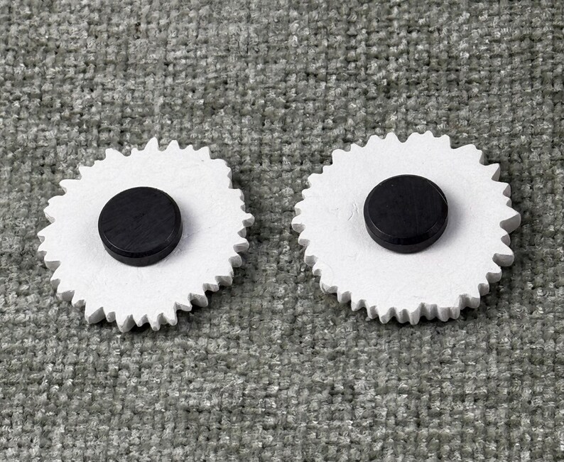 May include: Two white, gear-shaped magnets with a black, circular magnet in the center. The magnets are on a gray, textured surface. The gear-shaped design adds a unique touch to these functional magnets.