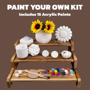 May include: A paint-your-own kit with the text "PAINT YOUR OWN KIT Includes 15 Acrylic Paints." The kit includes a wooden tiered display with various white ceramic shapes, sunflowers, paintbrushes, a palette, and small paint pots.