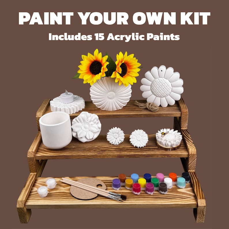 May include: A paint-your-own kit with the text "PAINT YOUR OWN KIT Includes 15 Acrylic Paints." The kit includes a wooden tiered display with various white ceramic shapes, sunflowers, paintbrushes, a palette, and small paint pots.