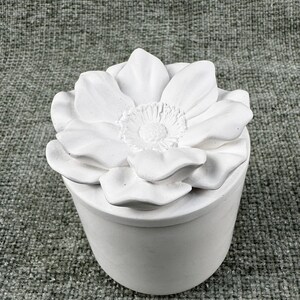May include: A white, cylindrical container with a lid shaped like a large flower. The flower has multiple petals and a detailed center. The container is made of a smooth, matte material.