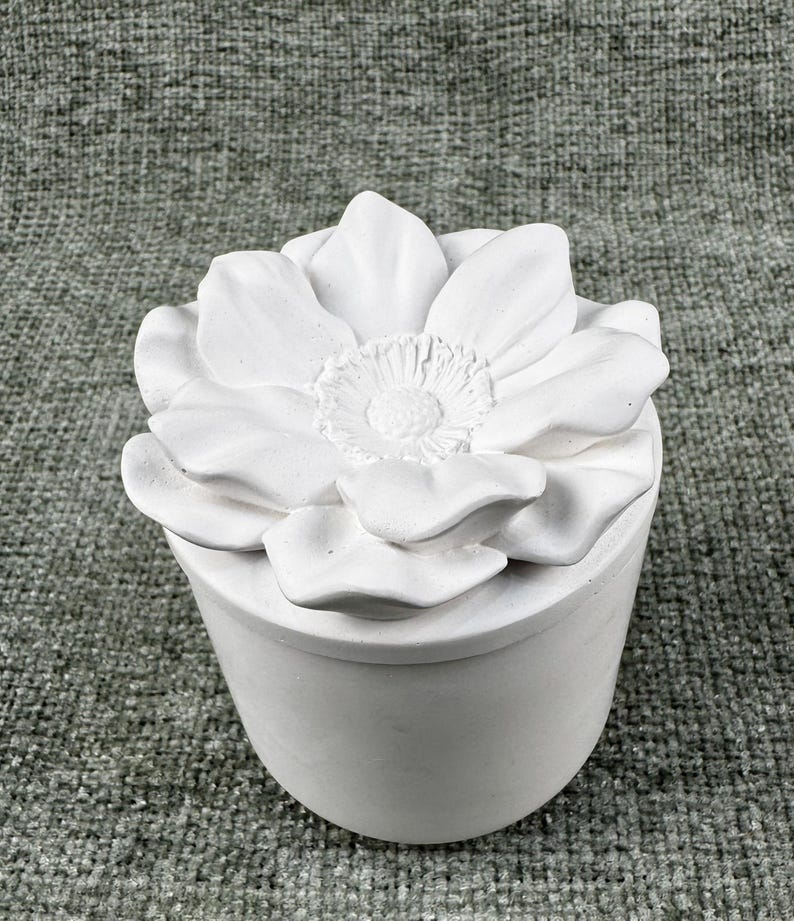 May include: A white, cylindrical container with a lid shaped like a large flower. The flower has multiple petals and a detailed center. The container is made of a smooth, matte material.