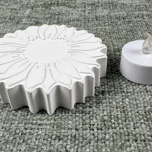 May include: A white, sunflower-shaped decorative piece with detailed petal carvings, next to a small, white LED candle. The candle has a realistic, flickering flame effect. The items are likely for home decor.