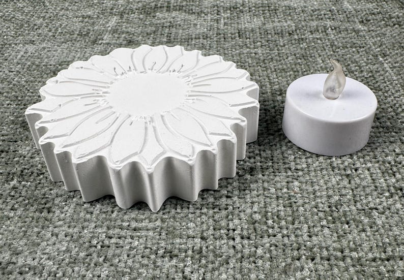 May include: A white, sunflower-shaped decorative piece with detailed petal carvings, next to a small, white LED candle. The candle has a realistic, flickering flame effect. The items are likely for home decor.