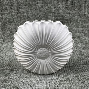 May include: A white, decorative, flower-shaped object with a textured surface. The object has a central, circular design surrounded by radiating, petal-like ridges. The piece is made of a solid, opaque material.