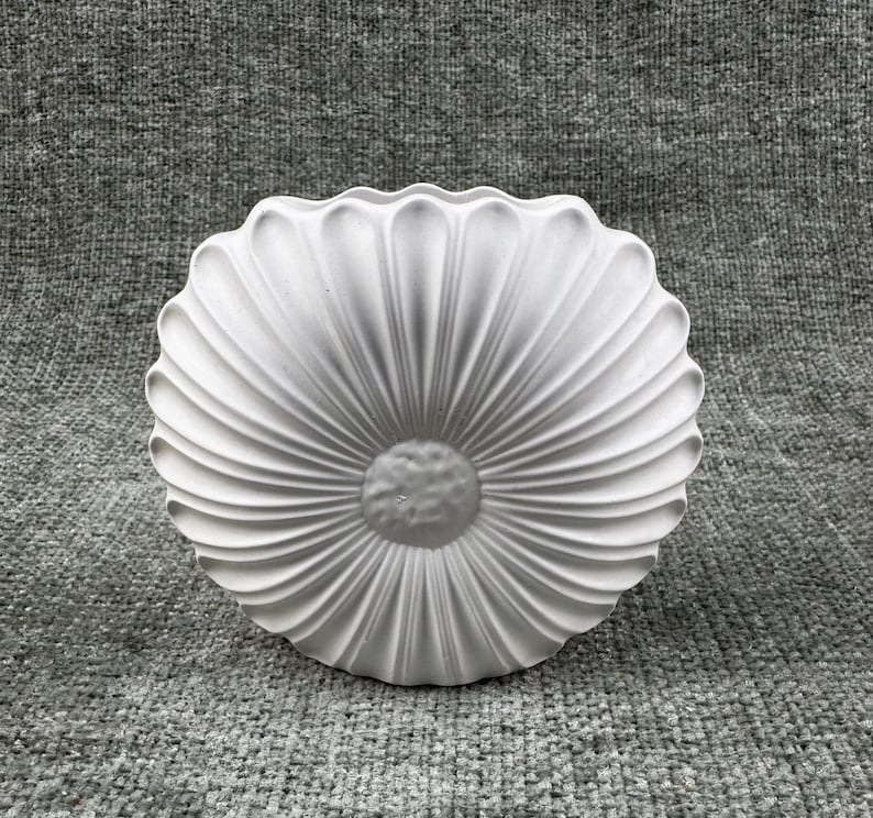 May include: A white, decorative, flower-shaped object with a textured surface. The object has a central, circular design surrounded by radiating, petal-like ridges. The piece is made of a solid, opaque material.