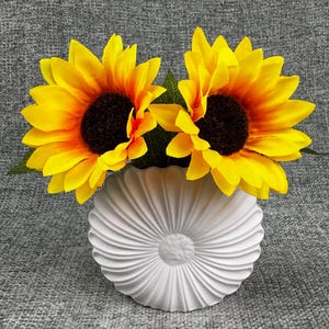 May include: Two bright yellow sunflowers with dark brown centers and green leaves are arranged in a white, textured vase. The vase has a circular shape with a ridged design, creating a floral aesthetic.