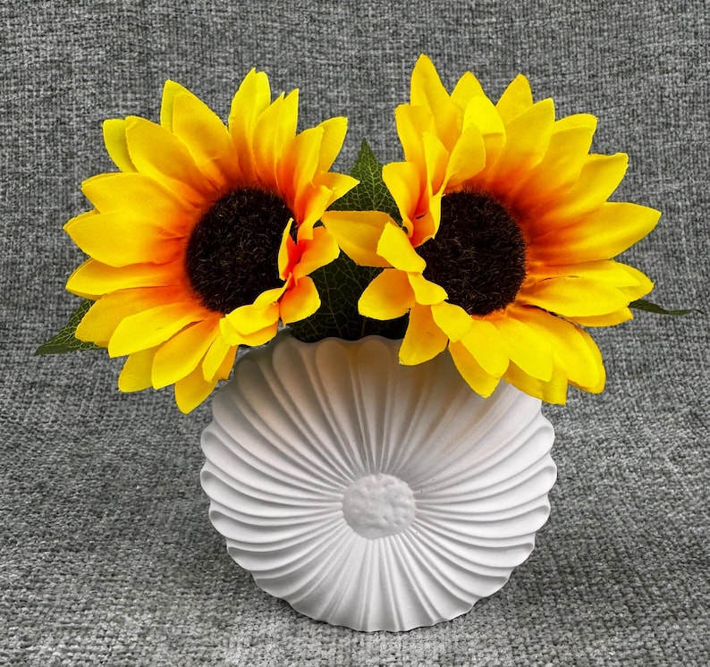 May include: Two bright yellow sunflowers with dark brown centers and green leaves are arranged in a white, textured vase. The vase has a circular shape with a ridged design, creating a floral aesthetic.