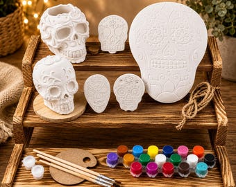 May include: A collection of white ceramic Day of the Dead skull decorations and painting supplies. The set includes various skull shapes, paint pots, paintbrushes, and a wooden palette, all arranged on a wooden shelf.