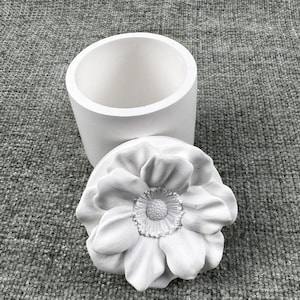 May include: A white, cylindrical container with a matching lid. The lid is shaped like a large, detailed flower. The container and lid are made of a smooth, matte material, likely ceramic or plaster.
