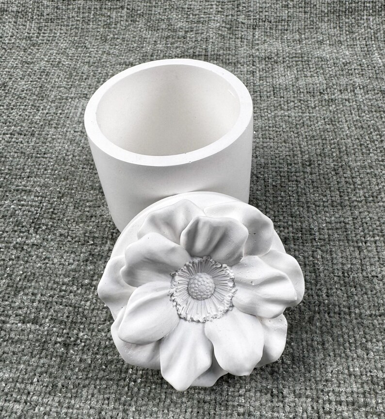 May include: A white, cylindrical container with a matching lid. The lid is shaped like a large, detailed flower. The container and lid are made of a smooth, matte material, likely ceramic or plaster.