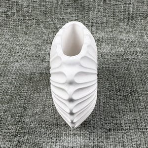 May include: A white, decorative vase with a textured, ridged design. The vase has an elongated, oval shape with a wide opening at the top. The surface has a series of curved, wave-like ridges that run vertically along the sides.