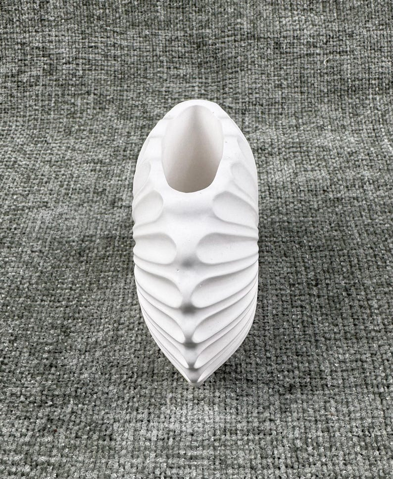 May include: A white, decorative vase with a textured, ridged design. The vase has an elongated, oval shape with a wide opening at the top. The surface has a series of curved, wave-like ridges that run vertically along the sides.