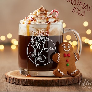 May include: A clear glass mug filled with a dark beverage, topped with whipped cream, caramel drizzle, and a candy cane. The mug has a floral design with the name "Josie". A gingerbread man stands beside the mug on a wooden coaster. Text reads "Christmas Gift Idea".