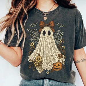 K&ouml;nnte beinhalten: Dunkelgraues T-Shirt mit einem Vintage-Geister-Design. Der Geist ist in Spitze geh&uuml;llt, mit einer braunen Schleife, Spinnweben, Flederm&auml;usen, Blumen und einem K&uuml;rbis. Ein Halloween-Grafik-T-Shirt.