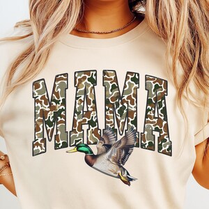 May include: Cream-colored t-shirt with the word "MAMA" in large, camouflage-patterned letters. A detailed illustration of a flying duck with green, brown, and white feathers is beneath the text. The shirt is a casual style.