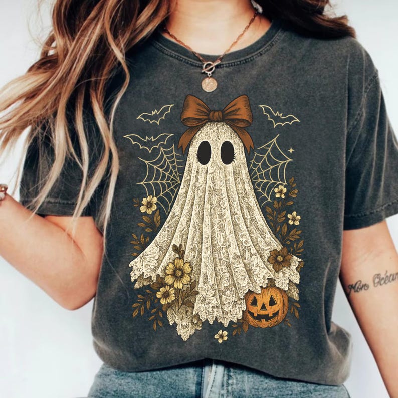 K&ouml;nnte beinhalten: Dunkelgraues T-Shirt mit einem Vintage-Geister-Design. Der Geist ist in einem spitzenartigen Stoff drapiert, mit einer braunen Schleife, Spinnweben, Flederm&auml;usen, Blumen und einem K&uuml;rbis. Ein Halloween-Grafik-T-Shirt.