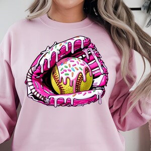 Puede incluir: Sudadera rosa con un gr&aacute;fico de un guante de softbol rosa y blanco con una pelota de softbol amarilla cubierta de glaseado blanco y chispas de colores.