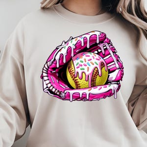 Puede incluir: Un guante de softball rosa y blanco con una pelota de softball amarilla en su interior. La pelota de softball est&aacute; cubierta de glaseado blanco y chispas de colores. El guante y la pelota de softball est&aacute;n goteando con glaseado rosa.