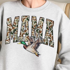 May include: A light gray sweatshirt with the word "MAMA" in a camouflage pattern. Below the text is a detailed illustration of a flying mallard duck with outstretched wings, showcasing brown, green, and yellow feathers.
