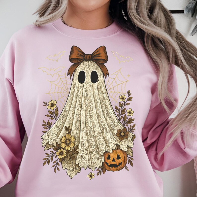 K&ouml;nnte beinhalten: Ein hellrosa Sweatshirt mit einem Halloween-Geist-Design. Der Geist hat eine braune Schleife, schwarze Augen und ein spitzenartiges Aussehen. Das Design umfasst einen K&uuml;rbis, Blumen und Spinnweben, wodurch eine festliche und gruselige &Auml;sthetik entsteht.