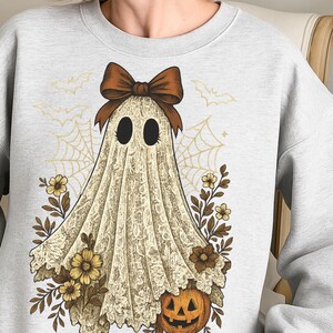 K&ouml;nnte beinhalten: Hellgraues Sweatshirt mit einem Geisterdesign im Vintage-Stil. Der Geist ist in einem spitzenartigen Stoff drapiert, mit einer braunen Schleife und floralen und K&uuml;rbis-Akzenten. Flederm&auml;use und Spinnweben vervollst&auml;ndigen das Halloween-Thema.